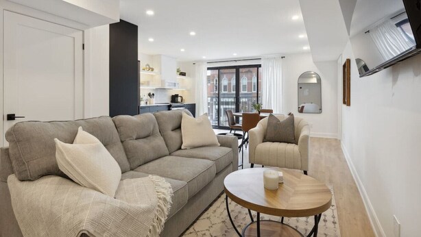 A modern living room with a sofa, armchairs, and a round wooden table. There is a kitchen area in the background with a dining table and chairs.