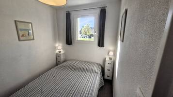 Une petite chambre à coucher, bien rangée, avec un lit simple, deux tables de chevet, une fenêtre avec des rideaux et un tableau encadré au mur.