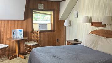 Un dormitorio con cama, una mesita pequeña con silla, una ventana con persianas y una lámpara fijada a la pared.