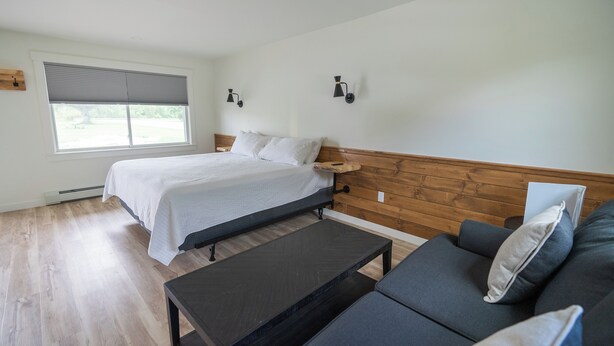 A bedroom with a bed, a wooden headboard, a dark-colored table, and a gray sofa. - Bingham Motor Inn (Bingham)