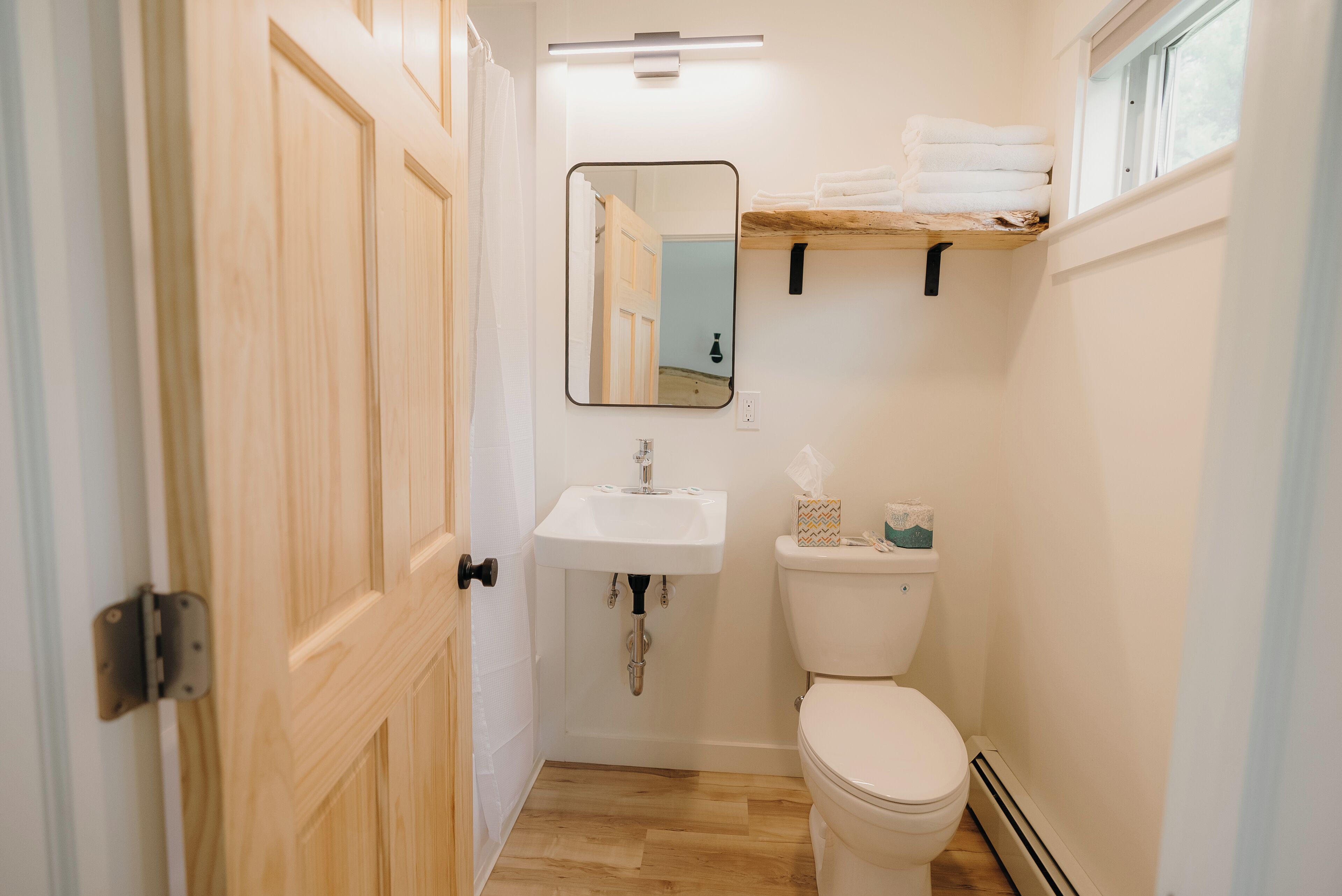 A small bathroom with a wooden door, a white sink, a toilet, and a mirror. There is a shelf with towels above the toilet.