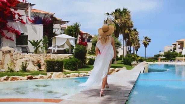 A woman in a white dress and straw hat walking along a poolside path with a pool and palm trees in the background. - Villa Elias 2BR Lower Apt with Pool (Maleme)