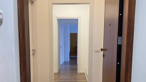 Egy folyosó, melynek padlója fából készült, a falai pedig fehérek, és amely egy ajtóval záruló szobába vezet.