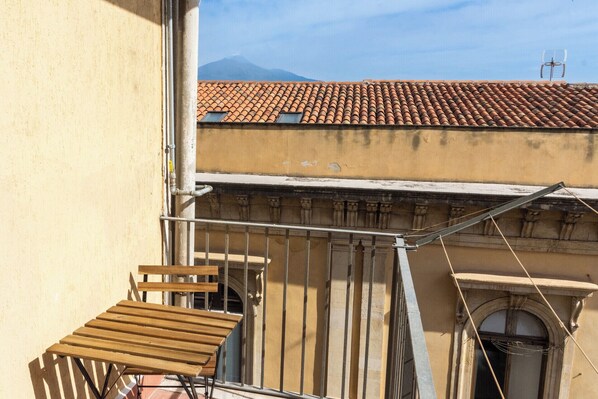 Un balcón con una silla de madera y baranda de metal que da a un edificio de techo a ladrillo y una montaña al fondo.