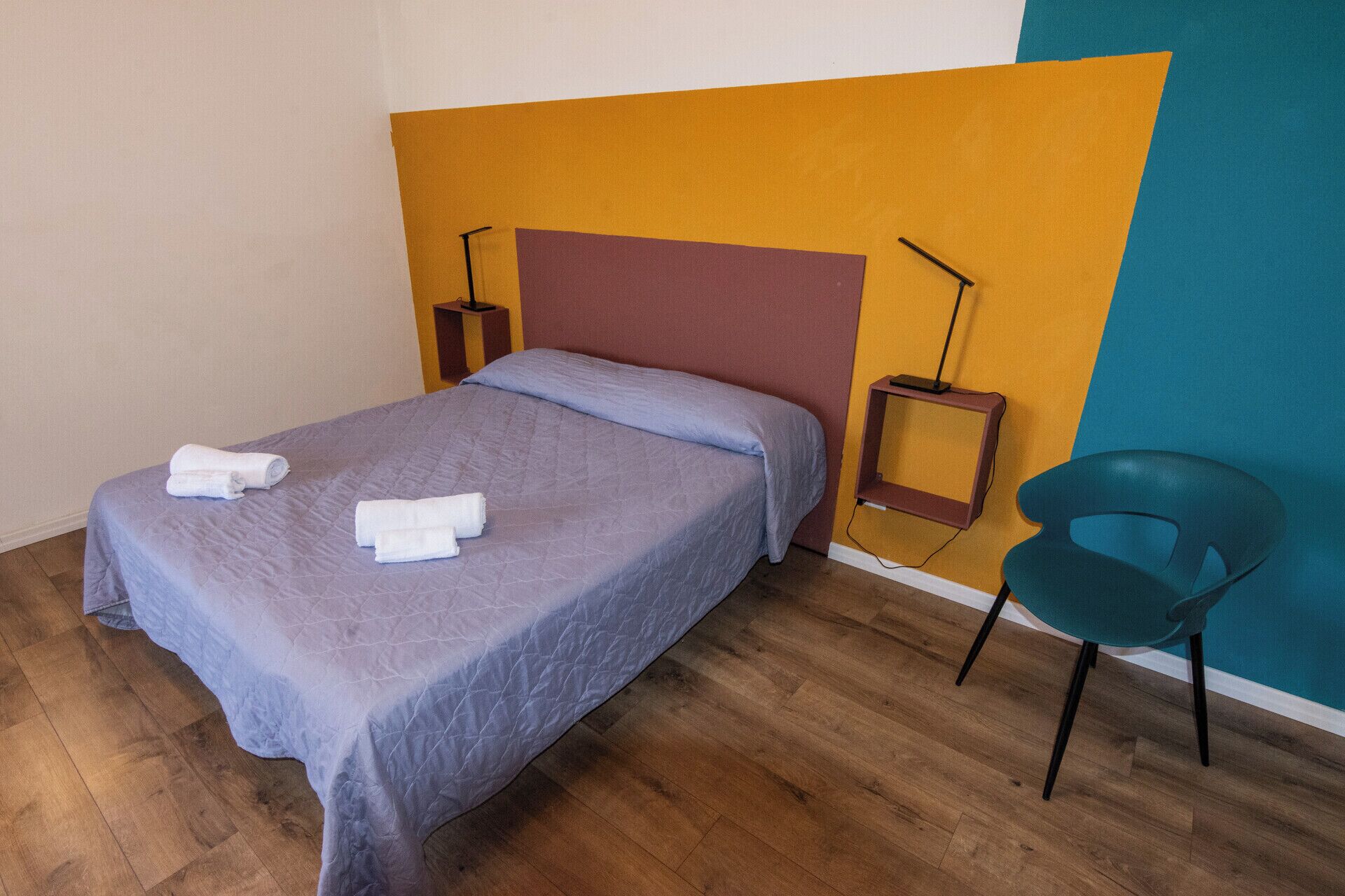 Un lit bien fait, recouvert d’une couvre-lit bleue et agrémenté de deux serviettes enroulées, une chaise de couleur turquoise et une table de chevet avec une lampe.