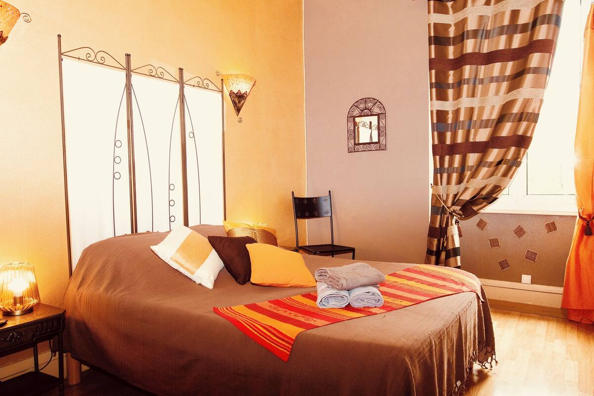 Una cama bien hecha con un plaid a rayas, una lámpara de noche y una mesita con lámpara.