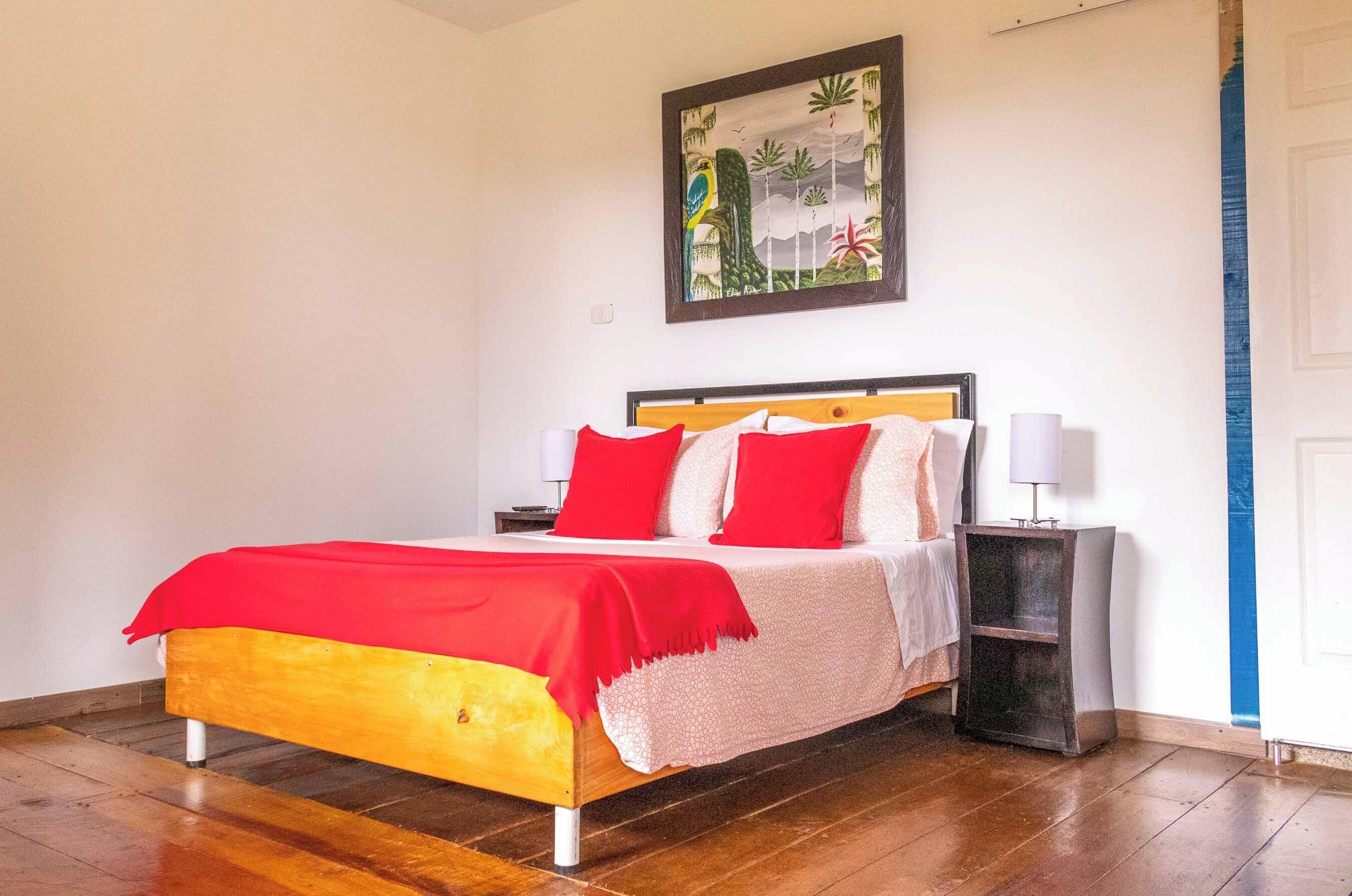 A bedroom with a wooden bed, red and white bedding, a nightstand with a lamp, and a painting on the wall.