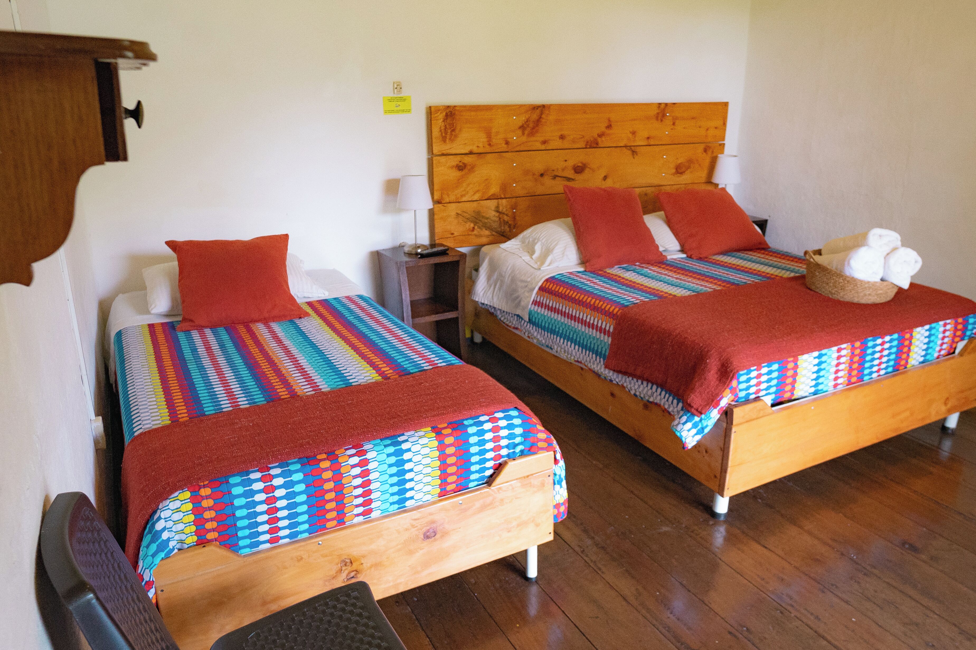 Deux lits en bois ornés de couvertures colorées et de coussins rouges, une table de chevet en bois avec une lampe, et un panier rempli de serviettes posé sur le sol.
