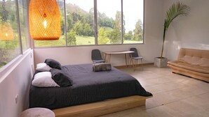 Ein modernes Schlafzimmer mit einem großen Bett, einem kleinen Tisch, zwei Stühlen, einer Couch und Blick auf eine grüne Landschaft durch große Fenster.