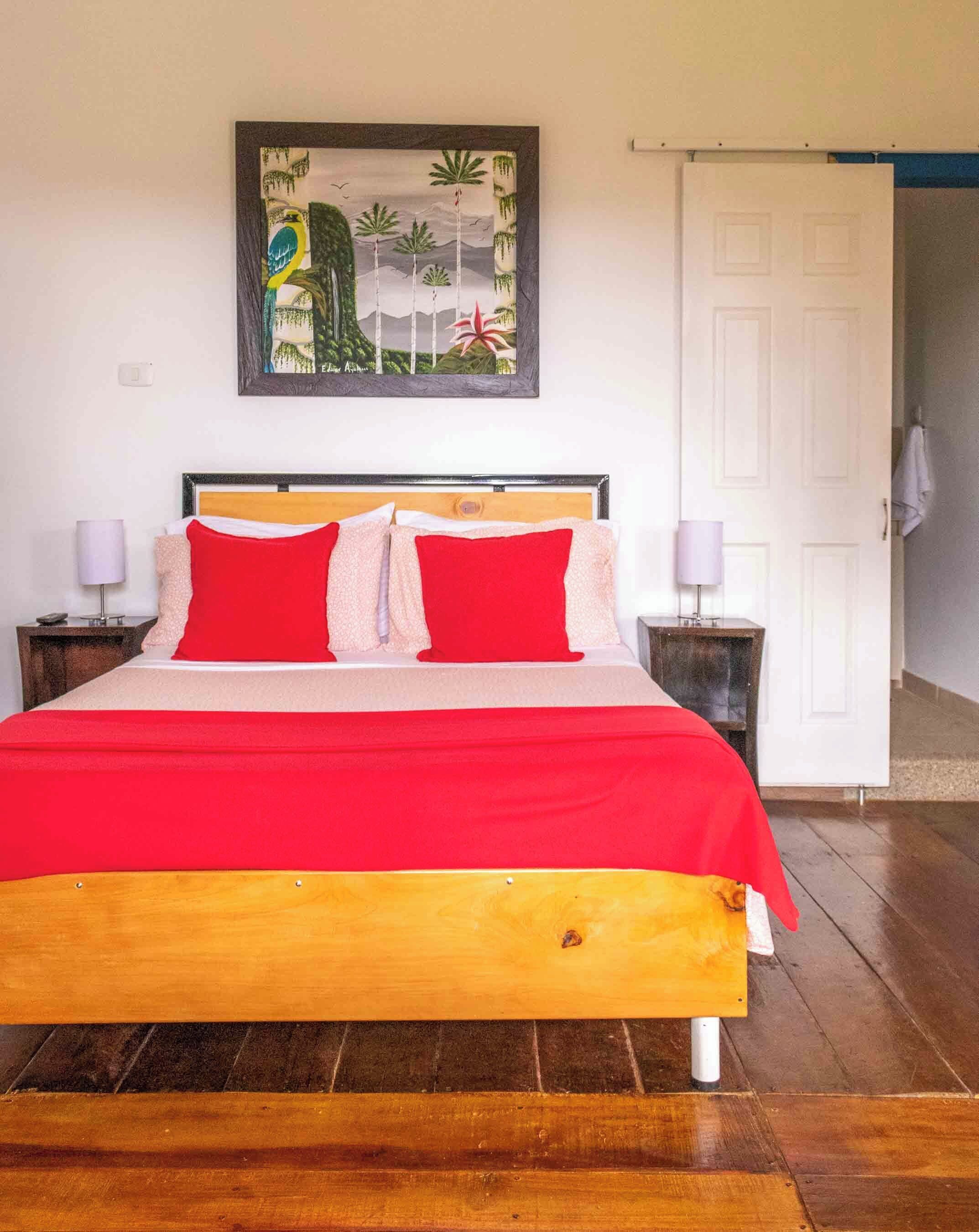 A bed with a wooden frame and red bedding, a nightstand with a lamp, a painting on the wall, and a white door.