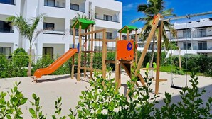 Un parque infantil con tobogán y estructuras para escalar, rodeado de vegetación y un edificio blanco con balcones.