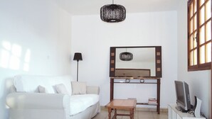 Ein Wohnzimmer mit einem weißen Sofa, einem Holztisch, einem Fernseher auf einem Schrank, einer Stehlampe und einem großen Spiegel über einer Konsole.