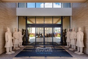 Una entrada moderna con puertas de vidrio, rodeada de estatuas de piedra, y un letrero que dice "Magic Village Views".