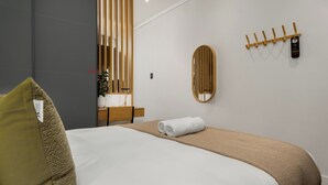 A modern hotel room with a neatly made bed, a wooden headboard, a bedside table with a lamp, a mirror, and a towel rack.