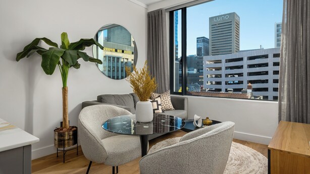 A modern living room with a round glass coffee table, grey sofas, a large potted plant, and a view of city buildings through large windows.