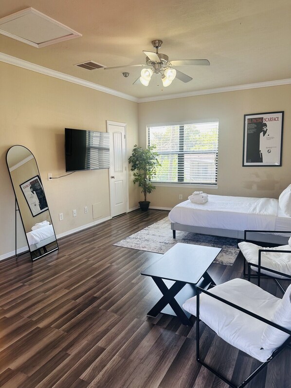 A bedroom with a bed, a desk, a chair, a TV, a ceiling fan, and a window with blinds.