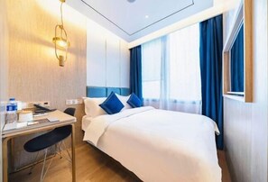 A hotel room with a bed, a desk, a chair, a desk lamp, a window with curtains, and a wall-mounted light fixture.