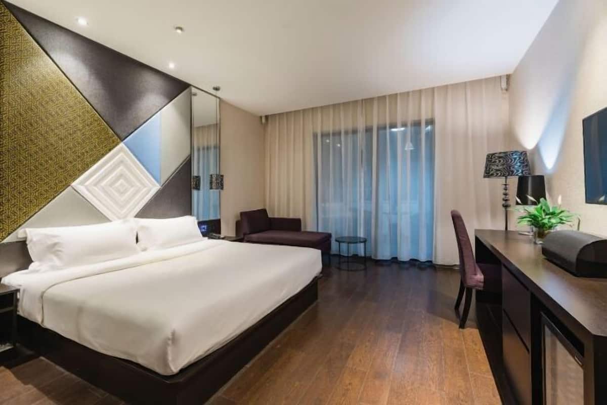 A hotel room with a large bed, a desk, a chair, a small table, and a large window with curtains.