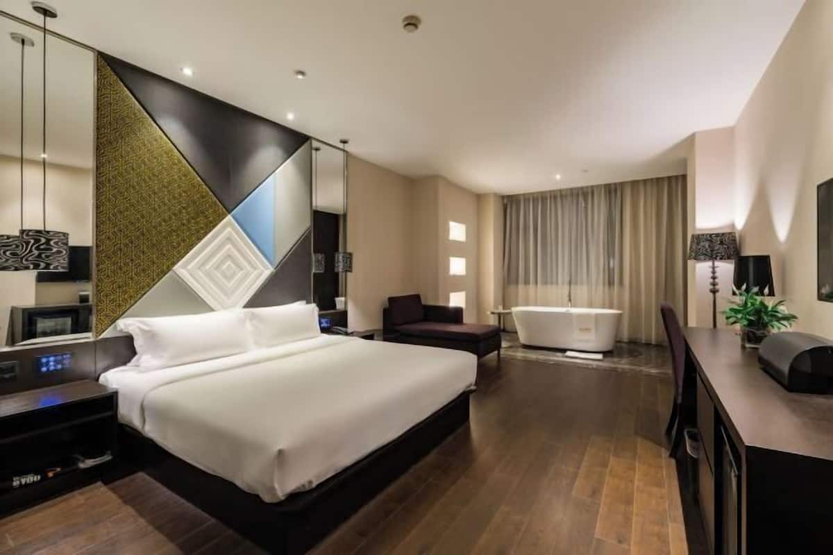 A modern hotel room with a large bed, a sitting area, a bathtub, and a desk with a lamp.