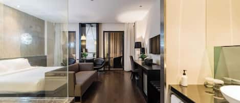 A modern hotel room with a large bed, a desk, a sofa, and a bathroom with a large mirror and sink.