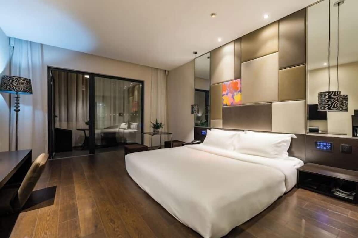 A modern hotel room with a large bed, a desk, a chair, a lamp, a mirror, and a sliding glass door leading to a balcony.