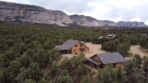 Una cabaña grande de troncos con una amplia terraza y una estructura más pequeña adosada, rodeada de un bosque denso y montañas al fondo.