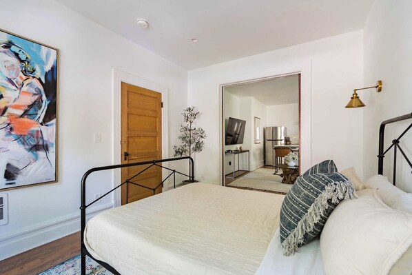 A bedroom with a bed, a wooden door, a painting, a plant, and a mirror reflecting a kitchen area.