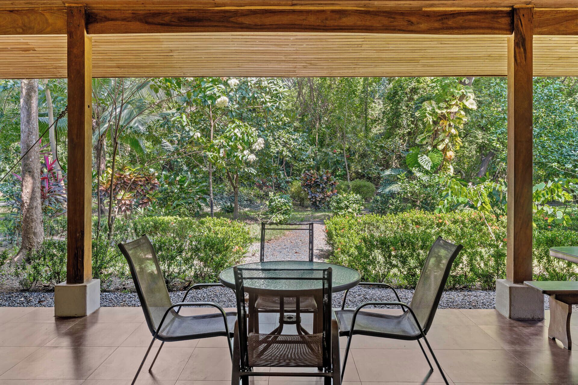 An outdoor patio with a round glass table and four chairs, surrounded by a wooden pergola and lush greenery.