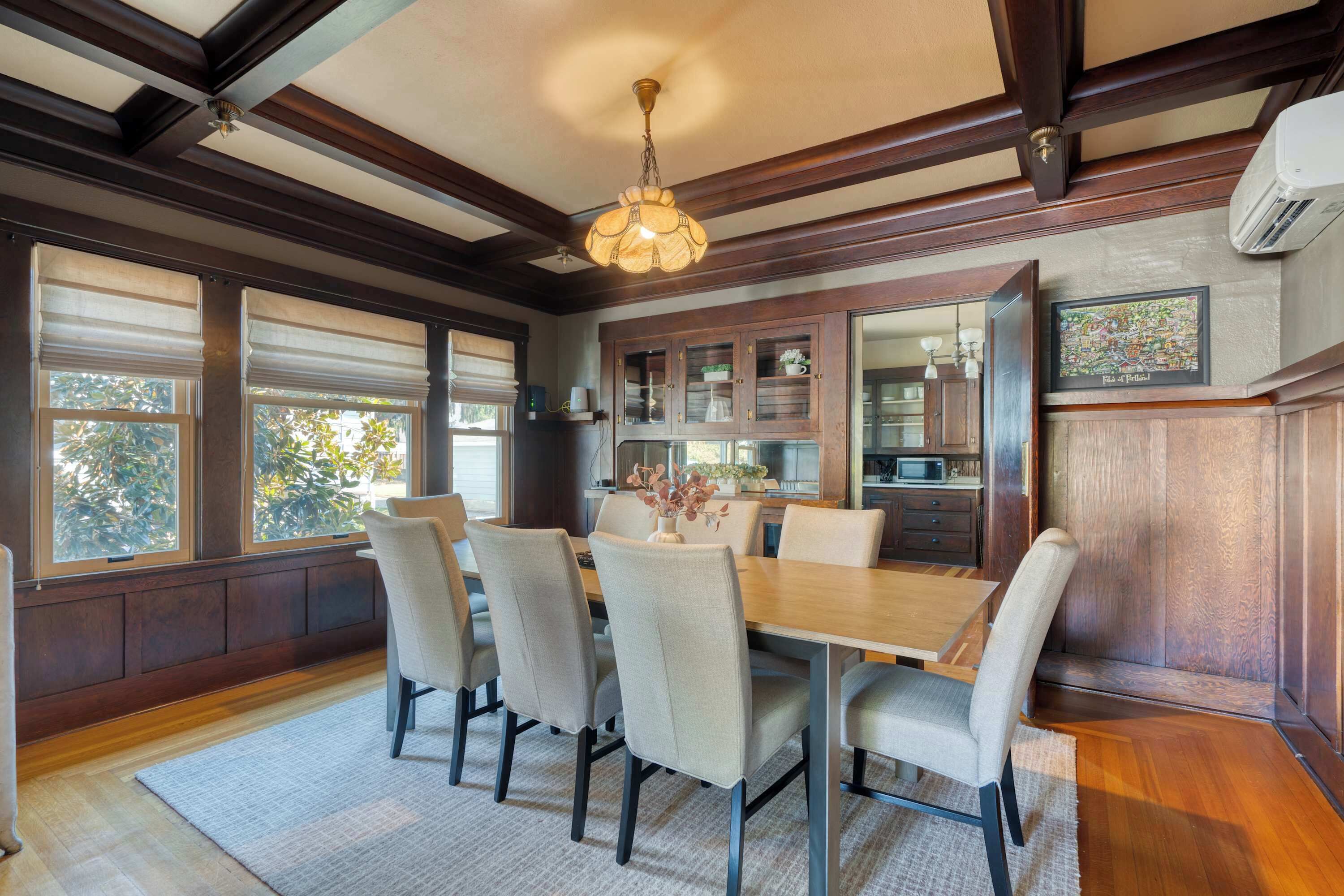 Un comedor con una mesa y sillas de madera, un candelabro y ventanas amplias con persianas.