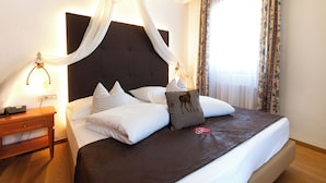 A neatly made bed with white linens and a brown headboard, a bedside table with a lamp, and a window with floral curtains. - Erlebnishotel Engel (Sluderno)