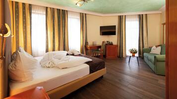 Quarto de hotel com uma cama, uma escrivaninha, uma televisão, um sofá verde e uma pequena mesa com uma planta.