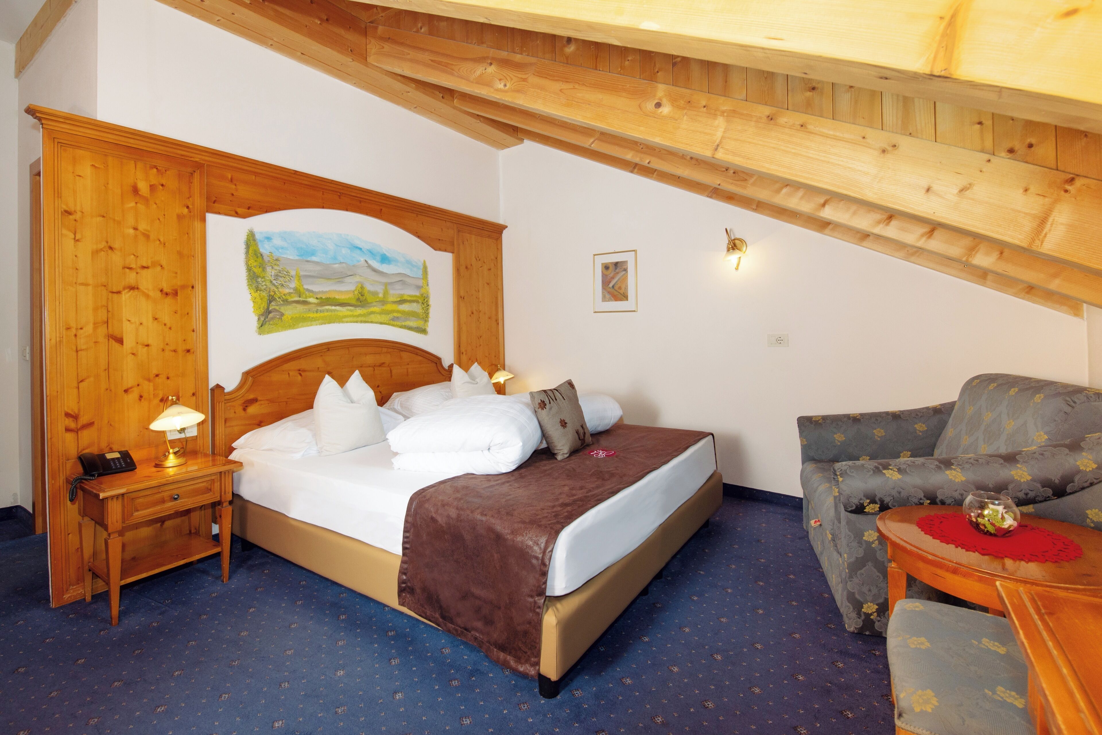 A bedroom with a bed, a nightstand, a wooden headboard, a painting, a sofa, and a small table with a red placemat.