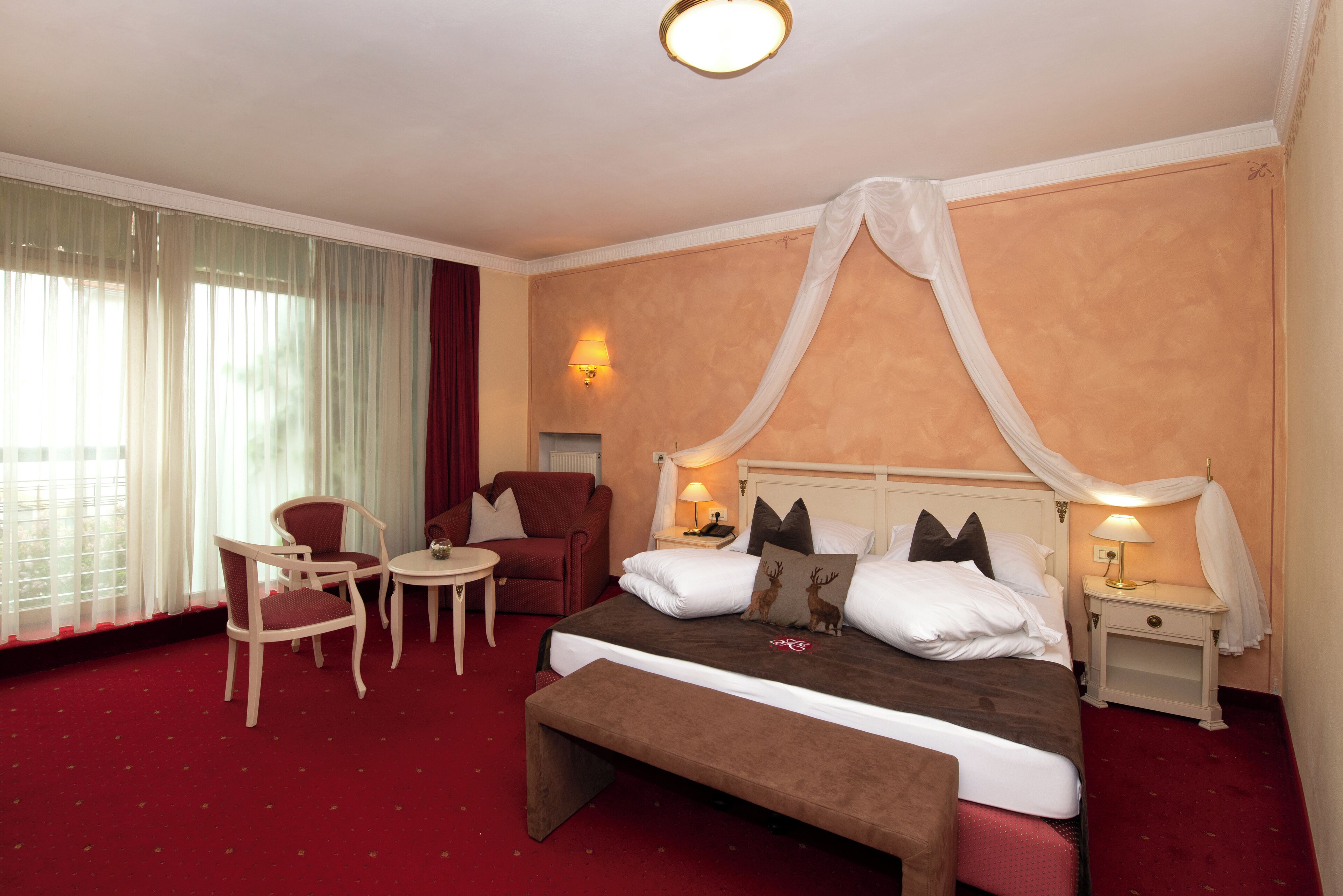 A hotel room with a large bed, a bench, two chairs, a small table, and a nightstand with a lamp.