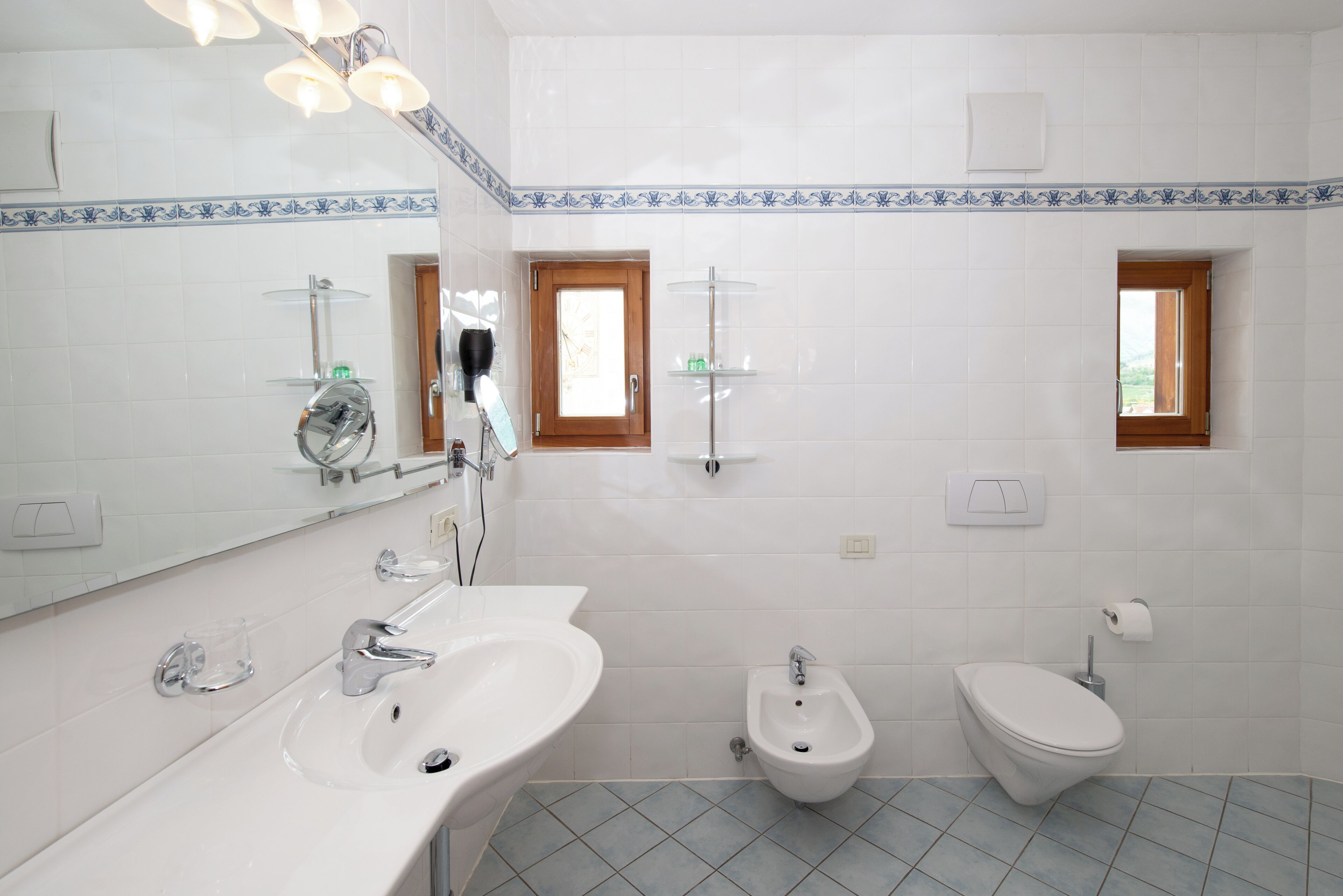 A bathroom with a sink, bidet, and toilet, tiled with white and blue patterns, and a mirror above the sink.