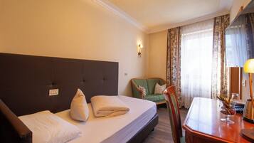 Ein Hotelzimmer mit Bett, einem grünen Sessel, einem Holztisch und einem Fenster mit Vorhängen.