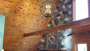 A cozy log cabin interior with a stone fireplace, wooden ceiling beams, and a large window. - Updated 3 bedroom A-Frame CABIN on the Fox River in Princeton with WiFi, AC (Princeton)