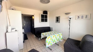 A compact living space with a sofa, a small table with a checkered tablecloth, a lamp, a refrigerator, and a door leading to another room.