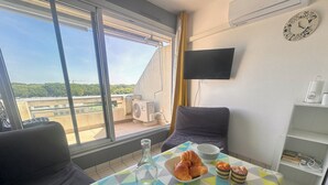A room with a table set for breakfast, including croissants, a bowl of cereal, and condiments. There is a TV mounted on the wall and a window with a view of greenery outside.