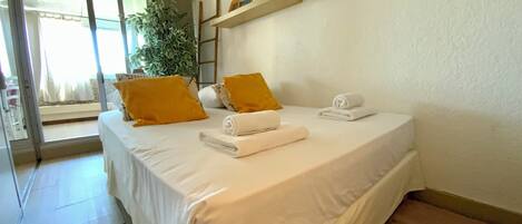 A hotel room with a bed, two pillows, a plant, a shelf with items, and a view through a glass door.