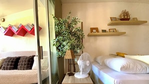 A bedroom with a bed, a small table with a fan, a potted plant, and shelves with decorative items.