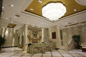 Eine Hotelhalle mit einer Rezeption, einer beeindruckenden Chandelüstere, Marmorböden und einer Treppe.