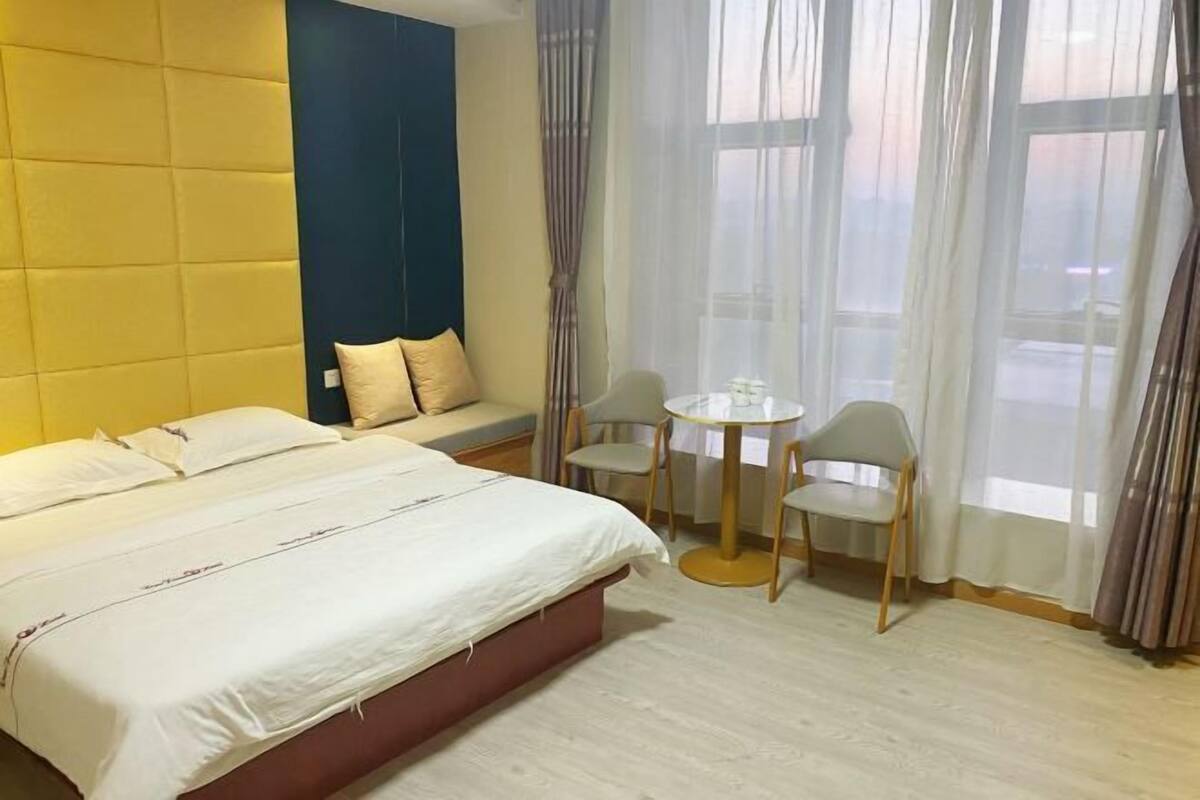Una habitación de hotel con cama, una mesita redonda pequeña, dos sillas, una ventana con cortinas y una pared azul decorativa.