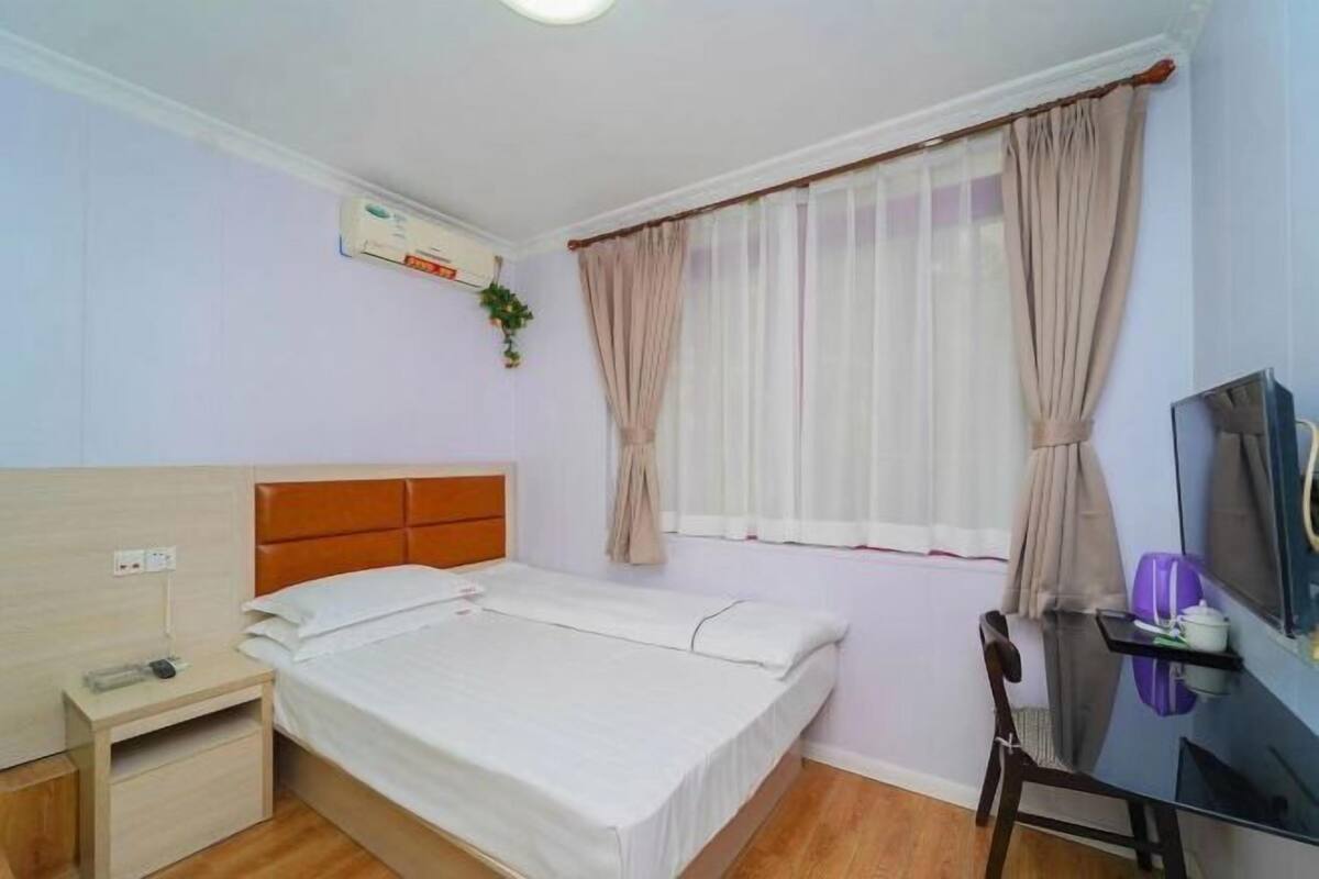 A hotel room with a single bed, a bedside table, a small desk with a chair, a TV, and a window with sheer curtains.