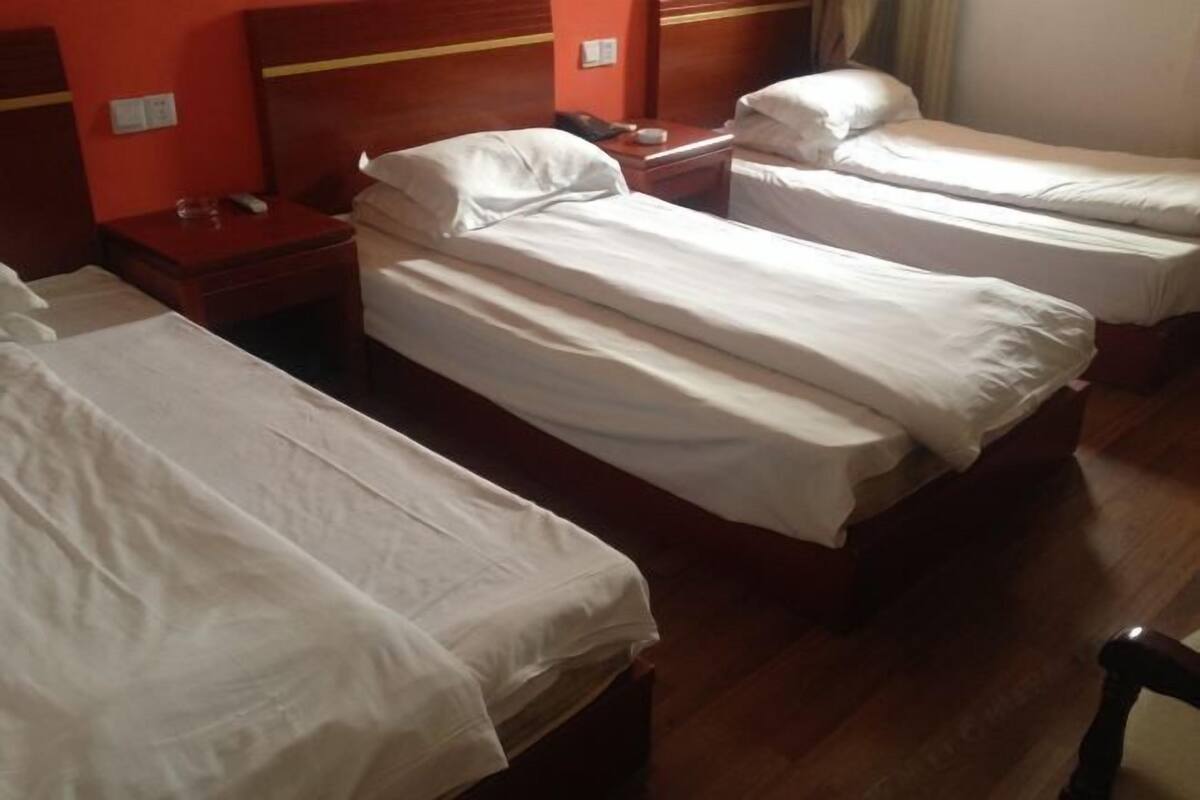 A hotel room with three single beds, wooden headboards, a wooden floor, and a small bedside table with a lamp and a phone.