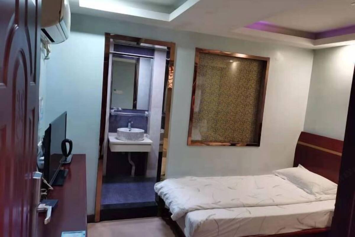 A hotel room with a bed, a bedside table, a mirror, a window with a patterned curtain, and a bathroom with a sink and mirror.