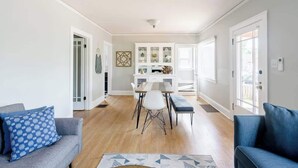 A modern living room with a dining area, white walls, and hardwood flooring. - Classic 1920s Alberta Home + 2 Tiny Homes (Portland)