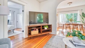 A modern living room with a flat-screen TV, wooden flooring, a dining area with chairs, and a large window providing natural light. - 2BR/1BA • Big Yard • Near Moda Center | Hot Tub (Portland)
