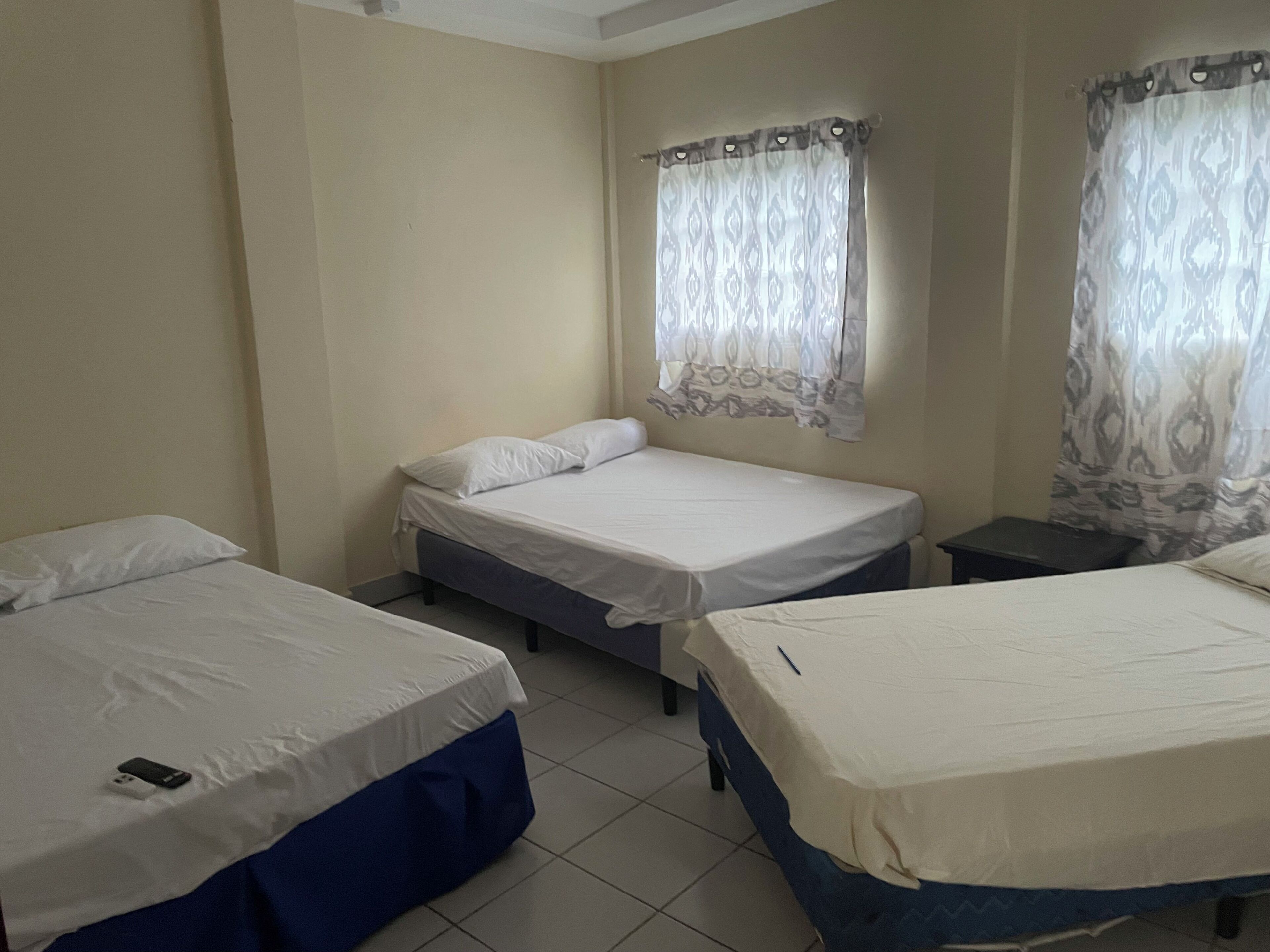 Habitación con tres camas individuales, cada una con colchón blanco y ropa de cama azul. Hay dos ventanas con cortinas estampadas, y las paredes están pintadas de un color claro.