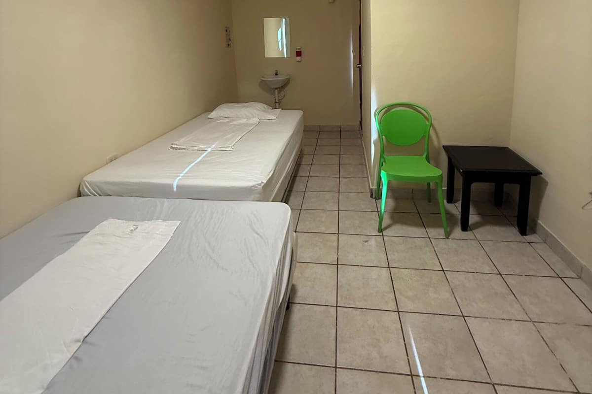 Una habitación pequeña y limpia con dos camas individuales, una silla verde, una mesita y un espejo en la pared.