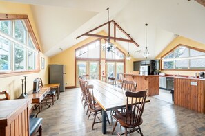 A spacious kitchen with a long dining table, wooden chairs, and a high ceiling with exposed beams.